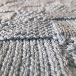 May include: Close-up of a light blue knitted fabric with a textured pattern. The fabric is soft and has a subtle sheen.