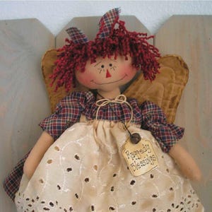 Puede incluir: Una muñeca de trapo hecha a mano con forma de ángel, con pelo de hilo rojo, un vestido a cuadros y alas color canela. La muñeca tiene una cara sonriente con rasgos cosidos y una etiqueta que dice "Raggedy Blessings". Es un artículo decorativo.