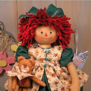 May include: A handmade rag doll with red yarn hair and a green bow. The doll wears a dress with a teddy bear pattern and holds a small teddy bear. The doll has painted facial features and is a charming, handcrafted item.