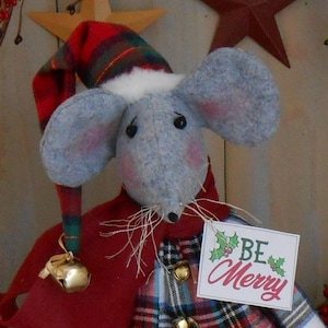 May include: A plush gray mouse doll wearing a red cape and a plaid hat with a white trim. The mouse has large ears, pink cheeks, and a tag that reads "Be Merry" with holly accents. A gold bell hangs from the cape.
