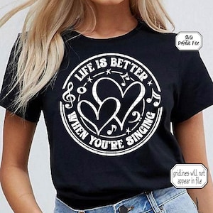 May include: Black t-shirt with a white circular design featuring the text "LIFE IS BETTER WHEN YOU'RE SINGING" around two heart shapes and musical notes. The shirt is short-sleeved and made of a soft material.