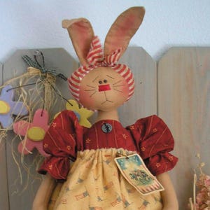 May include: A handmade Easter bunny doll with a painted face, red and white striped ears, and a red dress with star accents. The doll wears a yellow dress with American flag patterns and a tag.