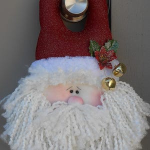 May include: A Santa Claus door hanger with a red glittery hat and a white yarn beard. The face is pink with small black eyes and a pink nose. Gold bells and festive greenery add to the holiday decoration.