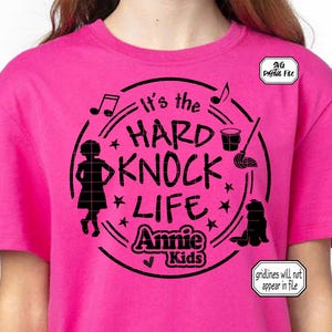 Könnte beinhalten: Ein leuchtend pinkfarbenes T-Shirt mit einem schwarzen Grafikdesign. Das Design zeigt den Text "It's the HARD KNOCK LIFE Annie Kids" in einem kreisförmigen Rahmen, zusammen mit Noten, Sternen und Silhouetten eines Mädchens, eines Hundes, eines Eimers und eines Wischmopps.