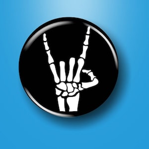 May include: A black and white button with a skeleton hand making the rock and roll hand gesture.