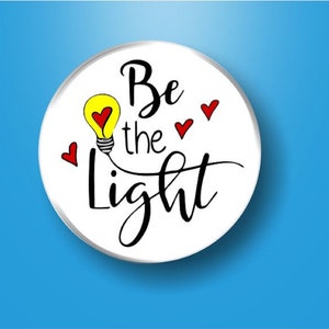 May include: White round button with the text "Be the Light" in black cursive font. A yellow light bulb with a red heart inside is drawn next to the text. Four red hearts are scattered around the text.