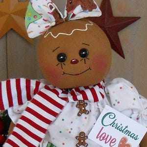 May include: A handcrafted gingerbread doll with a round head, rosy cheeks, and black button eyes. The doll wears a white dress with gingerbread man accents and a red and white striped scarf. A bow with a Christmas tree pattern adorns the head. A tag reads "Christmas love".