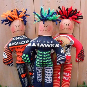 May include: Three handmade fabric dolls dressed in football team uniforms. One doll is in a Broncos uniform, another in a Seahawks uniform, and the third in a 49ers uniform. Each doll has a sad face and colorful yarn hair.