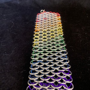 Pode incluir: Um bracelete de malha de corrente arco-íris com anéis prateados e detalhes de arame colorido. O bracelete é feito em um padrão de escamas e tem um fecho.