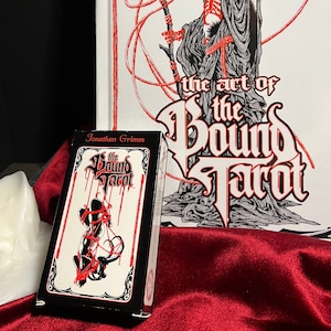 May include: A black box with red and white text that reads "The Bound Tarot" by Jonathan Grimm. The box is sitting on a red surface in front of a white book with black and red illustrations. The book cover reads "The Art of the Bound Tarot" by Jonathan Grimm.