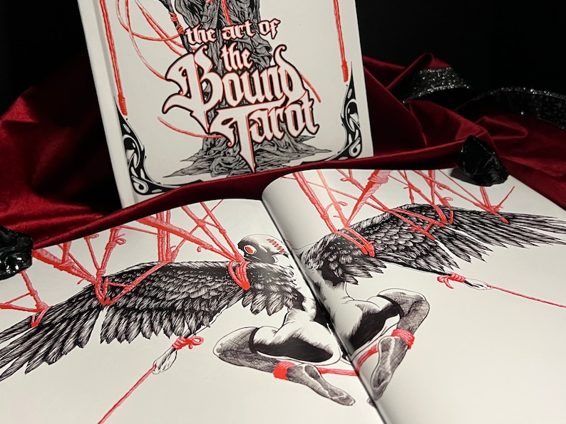 May include: Open book with the title "The Art of the Bound Tarot" on the cover. The book is open to a page with a black and white illustration of a figure with wings bound with red cords.