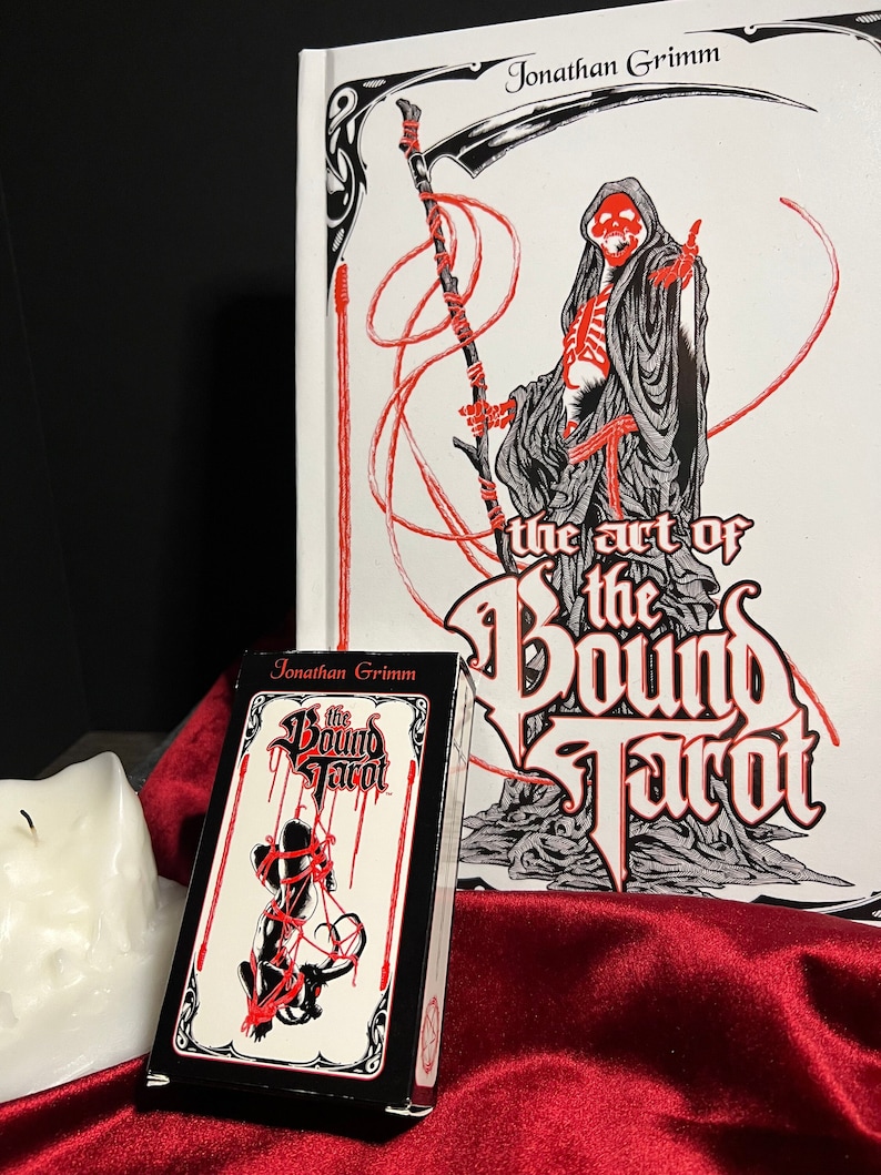 May include: A black box with red and white text that reads "The Bound Tarot" by Jonathan Grimm. The box is sitting on a red surface in front of a white book with black and red illustrations. The book cover reads "The Art of the Bound Tarot" by Jonathan Grimm.