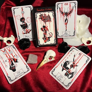 May include: A set of tarot cards with black and white illustrations of figures bound in red rope. The cards are laid out on a red velvet surface with a white candle, a bird skull, and black stones. The box for the cards is black with red text that reads "The Bound Tarot" and "Jonathan Grimm".