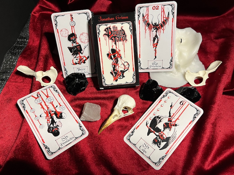 May include: A set of tarot cards with black and white illustrations of figures bound in red rope. The cards are laid out on a red velvet surface with a white candle, a bird skull, and black stones. The box for the cards is black with red text that reads "The Bound Tarot" and "Jonathan Grimm".