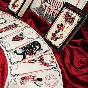May include: A set of tarot cards with red and black illustrations. The cards feature a variety of images, including swords, pentacles, and a figure with a gas mask. The cards are laid out on a red surface. The text on the cards reads "Three Swords", "Two Wands", and "Page of Wands".