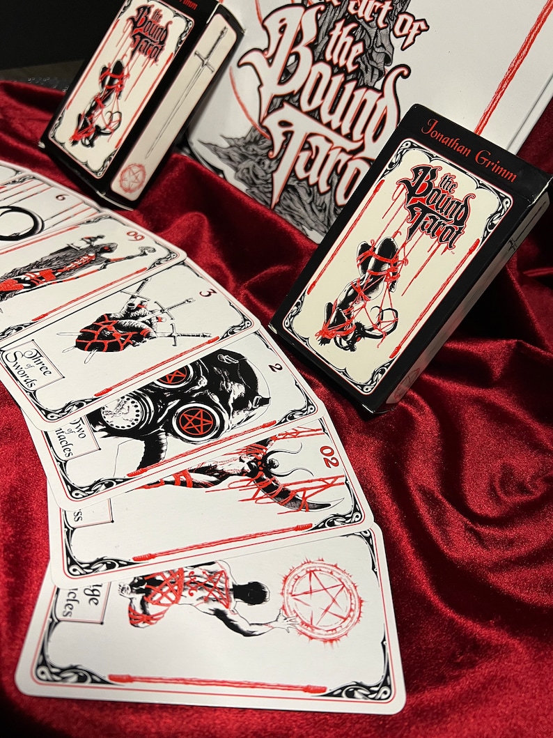 May include: A set of tarot cards with red and black illustrations. The cards feature a variety of images, including swords, pentacles, and a figure with a gas mask. The cards are laid out on a red surface. The text on the cards reads "Three Swords", "Two Wands", and "Page of Wands".