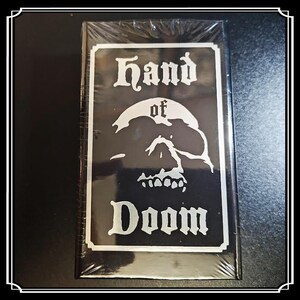 Op de afbeelding: Een zwart rechthoekig item met de woorden "Hand of Doom" en een schedelafbeelding in wit. Het item is verpakt in doorzichtig plastic. De tekst is in een gotisch lettertype.