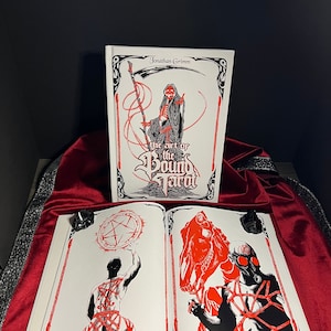 May include: A book titled "The Art of the Bound Tarot" by Jonathan Grimm. The cover features a skeletal figure in a hooded robe holding a scythe. The book is open to two pages, showing illustrations of the Page of Pentacles and the Knight of Pentacles.
