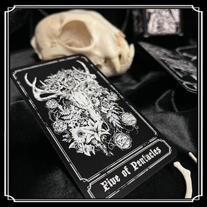 Peut inclure: Une carte de tarot noire avec une illustration blanche d'un cr&acirc;ne de cerf avec des fleurs et un pentagramme. La carte indique "Cinq de Pentacles".