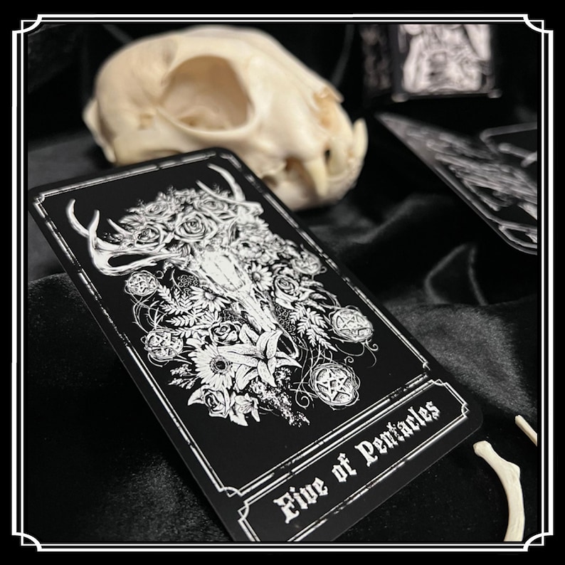 Peut inclure: Une carte de tarot noire avec une illustration blanche d'un cr&acirc;ne de cerf avec des fleurs et un pentagramme. La carte indique "Cinq de Pentacles".