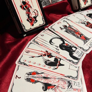 May include: A set of tarot cards with black and white illustrations and red accents. The cards feature a variety of figures and symbols, including a figure bound with ropes, a figure with a crown, and a figure with a sword. The cards are titled "The Bound Tarot".