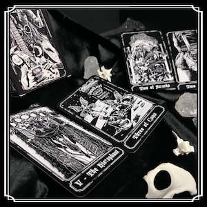 Peut inclure: Cartes de tarot noir et blanc avec des images gothiques et occultes. Les cartes pr&eacute;sentent des illustrations d'une figure squelettique avec une guitare, une figure avec une &eacute;p&eacute;e, une figure avec une coupe et une figure avec une couronne. Les cartes sont dispos&eacute;es sur une surface de velours noir.
