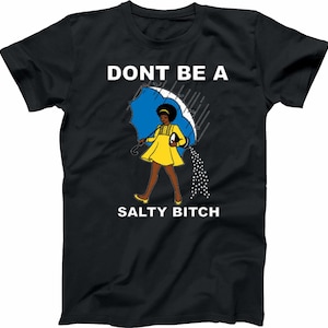 May include: Black t-shirt with the text "DONT BE A SALTY BITCH". The graphic features a woman in a yellow dress holding a blue umbrella, with salt pouring out of it. The design is a humorous statement.