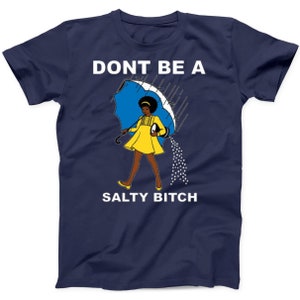May include: Navy blue t-shirt with the text "DONT BE A SALTY BITCH". The graphic features a cartoon woman in a yellow dress holding a blue umbrella, with salt pouring from it. A fun and bold statement tee.