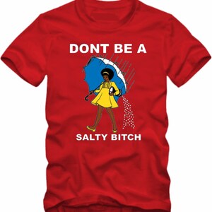 May include: Red t-shirt with the text "DONT BE A SALTY BITCH". The graphic features a cartoon woman in a yellow dress and holding a blue umbrella, with salt pouring from it. The shirt is a crew neck style.