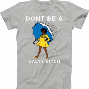 May include: A light gray t-shirt featuring a cartoon illustration of a woman in a yellow dress holding a blue umbrella. The text "DONT BE A SALTY BITCH" is printed above and below the illustration in white.
