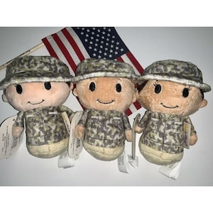 May include: Three plush toys dressed in camouflage military uniforms. The toys have brown eyes, rosy cheeks, and smiling faces. The toys are standing in front of a small American flag.