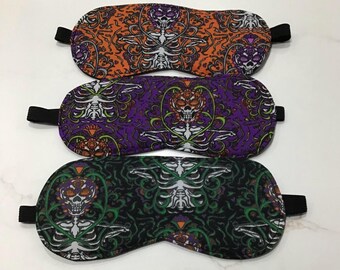 May include: Three sleep masks with black elastic straps. The masks display a skeleton design on orange, purple, and black backgrounds. The skeleton designs are surrounded by floral and decorative patterns. The masks are designed to block out light.