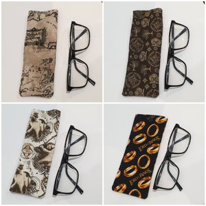 May include: Four different fabric eyeglass cases with a variety of designs. Each case is brown and features a different design. The designs include a map, a pattern of rings, a pattern of stars, and a pattern of rings. Each case is paired with a pair of black reading glasses.