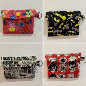 May include: Four different fabric coin purses with a flap closure and a metal ring. The purses are decorated with various patterns, including a pink floral pattern with a cartoon pig, a black and gray pattern with yellow bat silhouettes, a black and white newspaper print with the text "Dark Lord... More Sightings", and a red pattern with superhero faces.