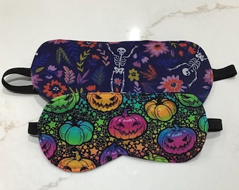 May include: Two sleep masks with black elastic straps. One mask features a dark blue floral and skeleton design. The other has a black background with a rainbow pumpkin design. Both masks are made of fabric.