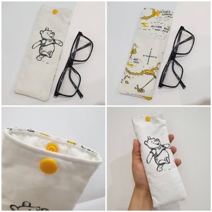May include: A white fabric glasses case with a Winnie the Pooh illustration and yellow button closures. The case is shown with a pair of black framed glasses inside.
