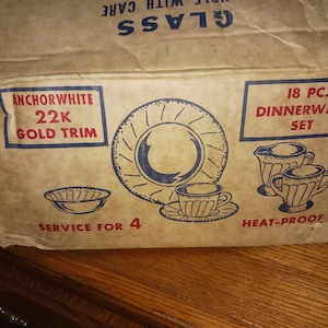 Op de afbeelding: Een vintage Anchorwhite 18-delige serviesdoos. De doos is bruin met blauwe en rode tekst en illustraties van borden, kopjes en een serveerkan. De doos leest "22K Gold Trim" en "Service for 4."