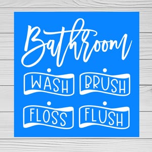 May include: A blue square sign with white text. The word "Bathroom" is written in a cursive font at the top. Below are four rectangular boxes with the words "WASH", "BRUSH", "FLOSS", and "FLUSH".