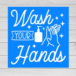 May include: A blue square sign with white text that reads "Wash Your Hands." The design includes a soap dispenser, hands, and starbursts. The sign is on a white wooden background.
