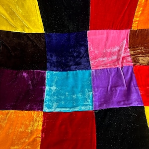 May include: A patchwork of colorful velvet squares in various shades, including yellow, black, red, pink, blue, purple, and orange. The fabric has a soft, textured appearance, with visible folds and creases. The arrangement creates a vibrant, geometric pattern.