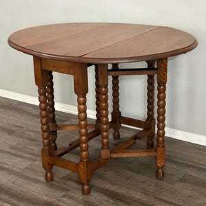 May include: A round, wooden drop-leaf table with a warm brown finish. The table features a turned-leg design with a bobbin-style pattern. The tabletop is split in the middle, allowing the leaves to be folded down. The table is approximately 30 inches in diameter.