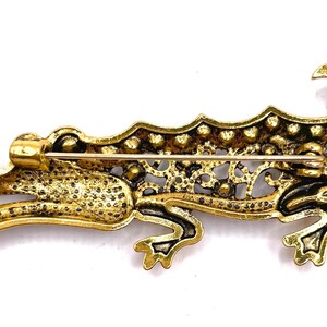 May include: A gold-toned alligator brooch with a detailed, filigree design. The brooch has a pin on the back for attaching to clothing.