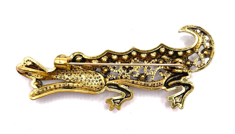 May include: A gold-toned alligator brooch with a detailed, filigree design. The brooch has a pin on the back for attaching to clothing.