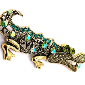 May include: A gold and green jeweled alligator brooch. The alligator is made of metal and has a detailed design. The alligator is decorated with green and clear rhinestones. The brooch is a decorative accessory.