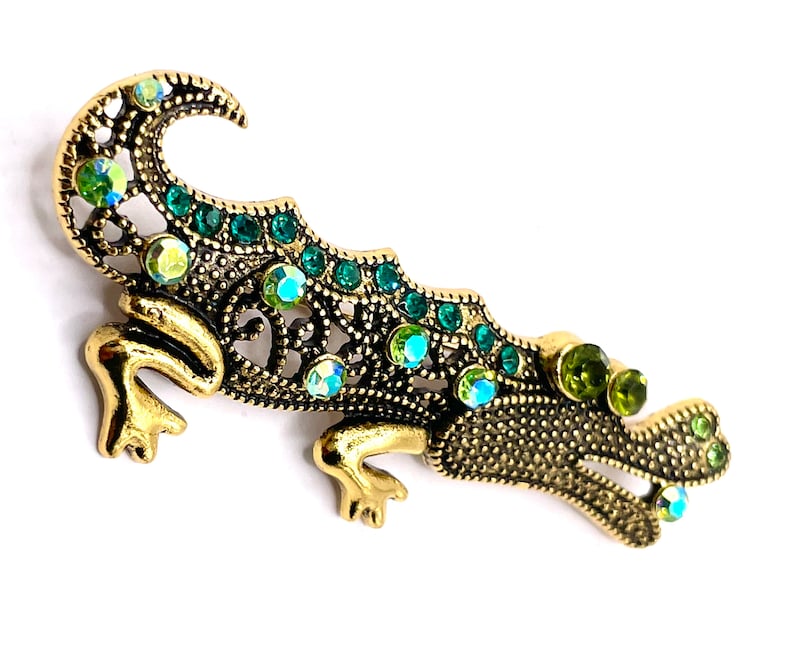 May include: A gold and green jeweled alligator brooch. The alligator is made of metal and has a detailed design. The alligator is decorated with green and clear rhinestones. The brooch is a decorative accessory.