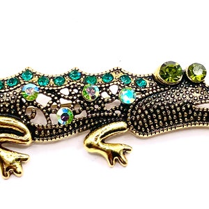 May include: A gold and green jeweled alligator brooch. The alligator is detailed with a textured body and has green rhinestones on its back and head.