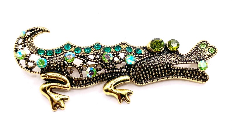 May include: A gold and green jeweled alligator brooch. The alligator is detailed with a textured body and has green rhinestones on its back and head.