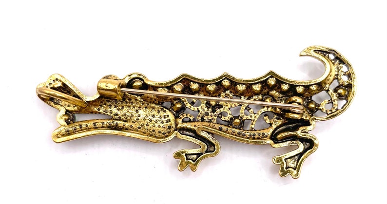 May include: A gold-toned alligator brooch with a filigree design and clear rhinestones. The brooch has a pin and catch closure on the back.
