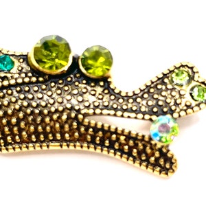 May include: A gold metal brooch in the shape of a stylized bird with green and iridescent rhinestones