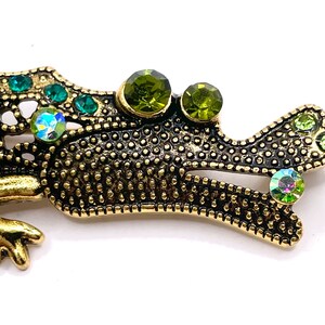 May include: A gold-toned metal alligator brooch with green and iridescent rhinestones. The alligator is in a walking position with its head turned to the side. The brooch is detailed with a textured pattern and has a shiny finish.